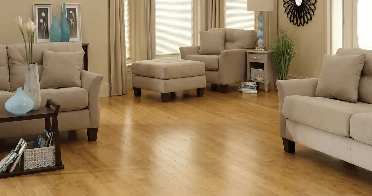 A living room with professionally installed bamboo flooring, beige sofas, a matching ottoman, a side table with a blue lamp, vases, framed pictures, and large windows with blinds and curtains in Cambridge, MA
