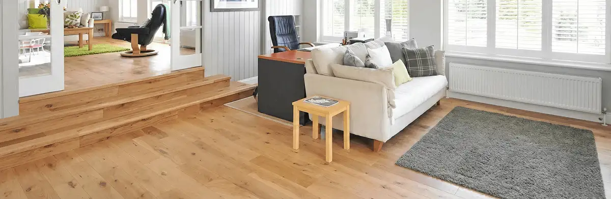 A modern living room with professionally installed laminate flooring, a beige sofa, a small side table, a gray rug, and large windows with white shutters in Cambridge, MA